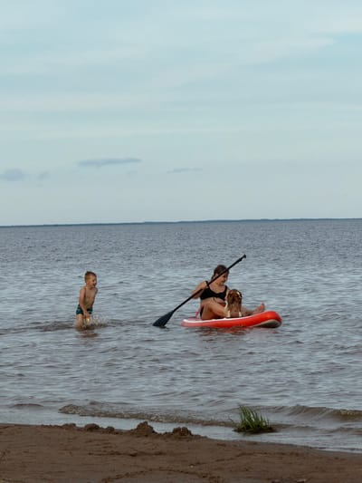 In a scene set in Ondvor, Russia, two people are engaged in water activities on a cloudy day. In the foreground, a young boy is splashing in the shallow water near the shore, wearing blue and green swim trunks. Further out in the water, a woman is seated on an orange paddleboard, holding a paddle. She is wearing a black swimsuit and appears to be interacting with a child or small dog also on the board. The water is choppy, indicating some wind, and the sky is overcast. The shoreline is sandy with some sparse vegetation visible. The overall mood seems relaxed and recreational, suggesting a summer day spent outdoors. There is no visible text in the image.