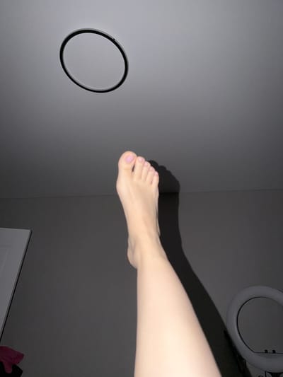 An indoor, low-light scene features a person's bare foot and lower leg, bathed in soft light, casting a distinct shadow on a grey wall. The foot, with lightly painted toenails, is extended upwards towards the ceiling. Above the foot, a circular object, possibly a ring light or a decorative fixture, is suspended from the ceiling. The background is composed of a neutral grey wall and a partial view of a white doorframe. The context of Razumnoye, Russia, is not discernible from the visual elements present in the image.
