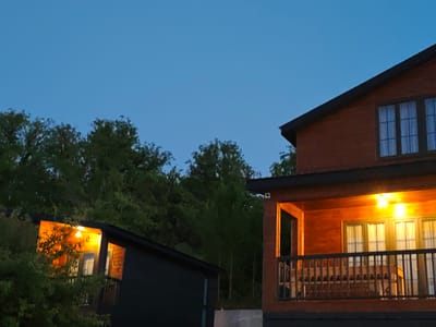 The image captures a twilight scene featuring two wooden structures nestled amongst lush green trees. On the right, a two-story wooden house with a dark roof is partially visible, showcasing an illuminated balcony with a wooden railing. Several chairs are arranged on the balcony, and bright interior lights are visible through the windows. To the left, a smaller dark-colored structure with a light illuminated from within is partially obscured by foliage. The sky above is a clear, deep blue, indicating the late hour. The location is identified as Ukeshi, Georgia.
