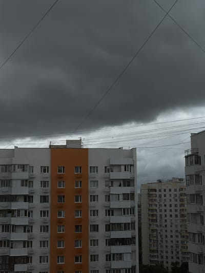 The image depicts a view of apartment buildings under a dark, overcast sky, suggesting inclement weather. Several power lines crisscross the upper portion of the frame, adding a sense of urban complexity. The buildings themselves are multi-story structures with numerous windows and balconies, characteristic of residential areas in Russian cities like Perepechino. One building features a striking orange facade section, contrasting with the predominantly white or grey exteriors of the surrounding structures. The scene is devoid of people, focusing entirely on the architectural and atmospheric elements. The overall impression is one of a mundane, everyday urban landscape under a somber sky.