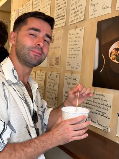 A man is seated at a wooden table, holding a paper cup with a wooden stirrer in his right hand. He wears a white and black patterned short-sleeved button-up shirt. The background consists of a wall covered with handwritten notes and a photograph. The man is looking towards the camera with a slight smile. One of the notes appears to be in Italian, mentioning "Bergamo" and "Mozzo, Italy." The setting suggests a casual establishment, possibly a cafe or restaurant, in Mozzo, Italy.