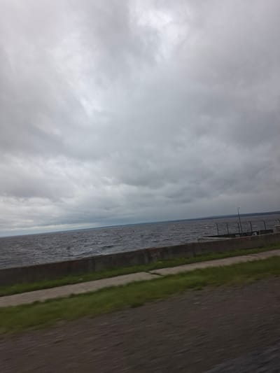 The image depicts a body of water with a cloudy sky overhead. The water appears choppy, suggesting it might be windy or that a boat has recently passed through. Along the right side of the image, there is a paved area with a strip of grass. A railing or fence runs along the edge of this paved area, parallel to the water. In the distance, the water stretches out to meet the horizon, which is obscured by the overcast sky. The overall impression is one of a somewhat gloomy or overcast day, possibly in a park or along a waterfront promenade. There is no visible text in the image. The crops provide closer views of the water, the cloudy sky, and the paved area with grass.