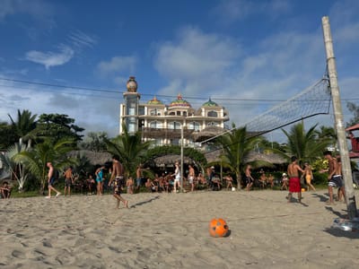 The image shows a beach volleyball game in progress in Montañita, Ecuador, located at coordinates -1.825, -80.754. The beach is sandy and the volleyball net is strung between two poles. There are about 20 people on the beach. Some are playing volleyball, while others are watching. There are palm trees and a building in the background. The building is a multi-story hotel with a large white facade and multiple domes. The scene is bathed in daylight and the sky is blue with scattered clouds. The air is clear and the beach is empty except for the people playing volleyball. The players are wearing swimsuits and shorts. Some are wearing hats. The players are having fun and enjoying the warm weather. The game is intense and the players are focused on winning. The volleyball is orange and white. It is sitting on the sand next to the net.