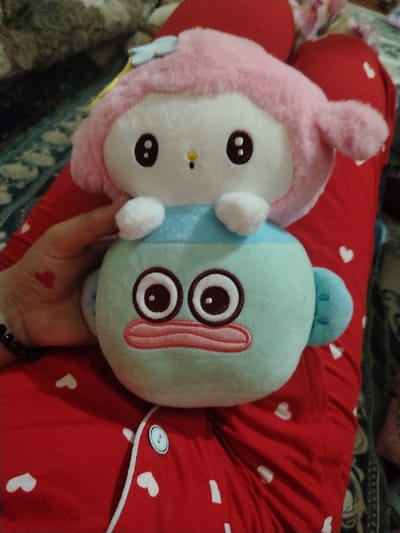 A person in red pajamas with white hearts holds two plush toys. The top one is pink and white, with dark, oval eyes and small hands. The bottom one is light blue with large, circular eyes and a wide, outlined mouth. The person's hand, adorned with a small red heart tattoo, supports the bottom toy. A bracelet is visible on the wrist. The pajamas have a button closure.