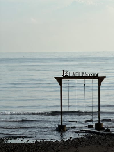 An outdoor, eye-level, medium shot captures a beach scene at the Labuan Resort in Pasir Putih, Indonesia. The image is dominated by the calm, blue ocean stretching to the horizon, with faint horizontal lines indicating gentle waves. A wooden swing set with two swings, partially submerged in the water, stands prominently in the foreground. Above the swings, a metallic sign reads "LABUAN RESORT." The shoreline is covered with dark pebbles and some water. The sky above is a pale blue with a few wispy clouds. The lighting suggests it is either early morning or late afternoon. There are no people visible in the image.