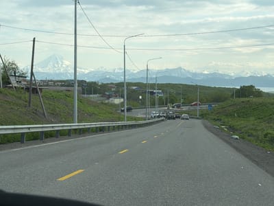 The image depicts a scenic view of a road in Krutoberegovy, Russia, leading towards a majestic mountain range capped with snow. The road, marked with yellow lines, curves gently to the right, with a metal guardrail on the left side. Several vehicles, including cars and SUVs, are driving on the road, suggesting a moderate level of traffic. The surrounding landscape is characterized by lush green hills and scattered trees, with a few buildings visible in the distance. The sky is overcast with clouds, indicating a possibly cool or temperate day. In the background, the imposing, snow-covered mountains create a dramatic backdrop, with a hint of the sea or a large body of water visible on the far right. The overall scene conveys a sense of natural beauty and rural tranquility, with hints of human presence through the road and vehicles. The time of day appears to be daytime, given the natural light.