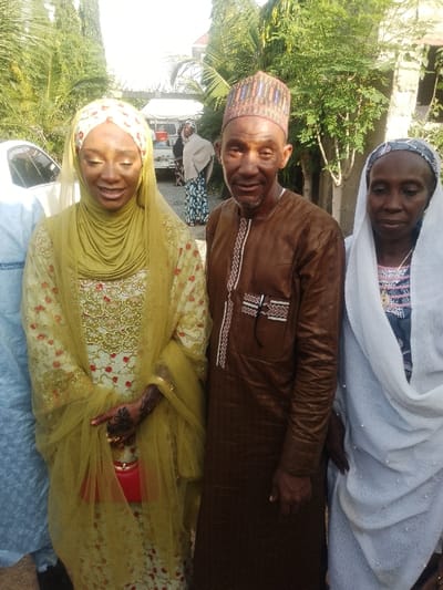 This image, taken in Unguwa Uku Kauyen Alu, Nigeria, displays three individuals posing for a photograph in an outdoor setting under bright daylight. On the left, a young woman is observed smiling, wearing a cream-colored long-sleeved dress with intricate gold and red embroidery, a chartreuse green hijab, and a sheer green veil. Her left hand exhibits dark henna patterns and holds a small red clutch. Adjacent to her, a man with a light beard wears a brown long-sleeved tunic (kaftan style) featuring white embroidered patterns on the chest and cuffs, along with a multi-colored patterned cap; he looks directly towards the camera with a subtle smile. To the man's right, an older woman is visible, dressed in a patterned light blue garment and a white hijab adorned with small beads. A partial figure in light blue clothing is visible on the far left. The background consists of abundant green foliage, including palm trees and bushes. A white utility vehicle with red containers in its bed is parked in the mid-ground on the left, near two additional figures. A building structure is partially visible on the far right.