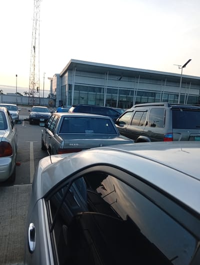 The image captures a parking lot scene in Omagwa, Nigeria. A mix of parked cars, including sedans and an SUV, occupy the foreground. Notably, one car has "OLVO" as branding on its rear. In the background, a modern-looking building with large windows is visible, possibly an airport terminal. A tall telecommunications tower stands adjacent to the building, with two workers visible ascending the tower. The sky suggests either early morning or late afternoon lighting.
