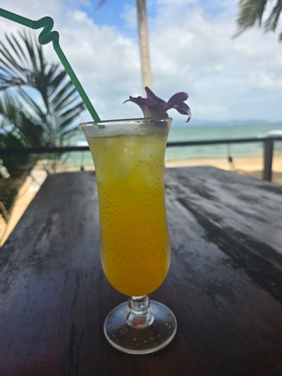 A hurricane glass filled with a yellow beverage, adorned with a purple orchid and a green straw, rests on a dark wooden surface. In the background, a blurred view of a beach with the ocean and sky is visible. A palm tree is partially visible in the lower left corner. The location is likely near the beach in the city of Ban Khlong Hia, Thailand.