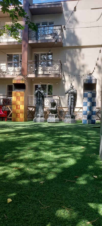The image displays a decorative outdoor setting with a focus on a chess-themed installation. In the foreground, artificial turf covers the ground, dappled with the shadows of overhead foliage. To the left, a building with cream-colored walls features several windows with decorative ironwork. A string of lights with exposed bulbs is strung across the scene.
The central elements are large, dark statues resembling chess pieces, specifically king and queen figures. These statues are positioned on pedestals, with one in the foreground and another further back. Behind the statues, a tall, checkered structure with blue and white patterns is visible, reminiscent of a rook or a tower in chess. Further to the right, another chess piece statue is partially visible. Below the statues, a section of the building's facade is decorated with a repeating pattern of yellow and red squares, each containing a stylized animal emblem, potentially indicating a connection to a specific house or team.
The scene appears to be outdoors, likely in a courtyard or garden area. The lighting suggests it is daytime, with the sun casting strong shadows. The presence of the chess pieces and decorative elements implies a deliberate design for aesthetic or recreational purposes, possibly a themed attraction or a public art installation. No people are visible in the image. The overall impression is one of a whimsical and visually engaging outdoor space.