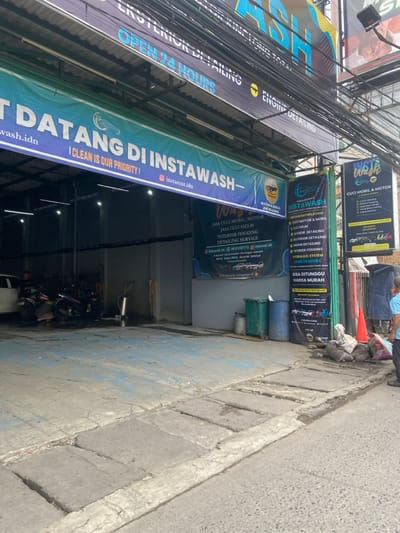 The image shows the exterior of a car wash in Bojongsoang, Indonesia, located at coordinates -6.972, 107.636. The car wash is called "Instawash" and features a large sign with the slogan "Datang di Instawash - I Clean is Our Priority!". The sign is in Indonesian and English. The car wash appears to be open 24 hours a day. There is a smaller sign next to the main one advertising various services including car and motorcycle washing, detailing, fogging, and cleaning. The building has a large open doorway that leads to the car wash area. There is a person in the doorway, but their face is not visible. The person appears to be wearing a light blue shirt. There are several motorcycles parked in the car wash area. A green trash can with a black trash bag is visible in the image. There is a person standing to the right of the trash can, wearing a blue shirt. There is a small orange traffic cone nearby. The ground in front of the car wash is paved with concrete. There are signs with a yellow background and black letters. The signs mention various services offered by the car wash. The image is taken during the day, and there is no sign of rain. There is a building behind the car wash with a "ASH" sign on it.