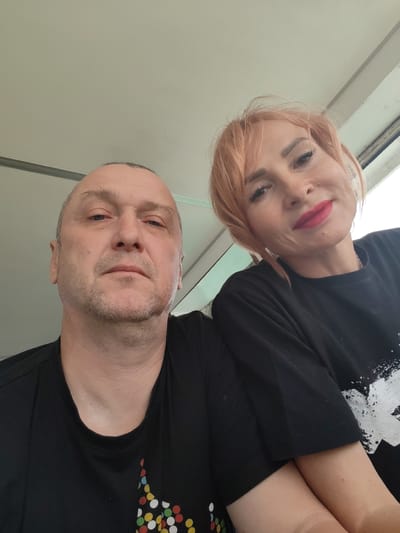 This is a selfie of a man and a woman. The man is in the foreground, on the left side of the frame, and the woman is behind him and to his right. Both are wearing black t-shirts. The man has a shaved head with some grey hair around the sides and a light stubble on his face. His expression is neutral. The woman has short, peachy-pink hair and is wearing bright red lipstick. She is smiling faintly, with her eyes looking slightly to her right.
The scene appears to be indoors, with a light-colored ceiling and wall visible behind them. A window is partially visible on the right side of the frame, suggesting the setting might be a room with a view, possibly during daylight hours, though no specific weather conditions can be determined. The overall lighting is soft. There is no visible text in the image. The subjects appear relaxed and are posing for a casual photograph.