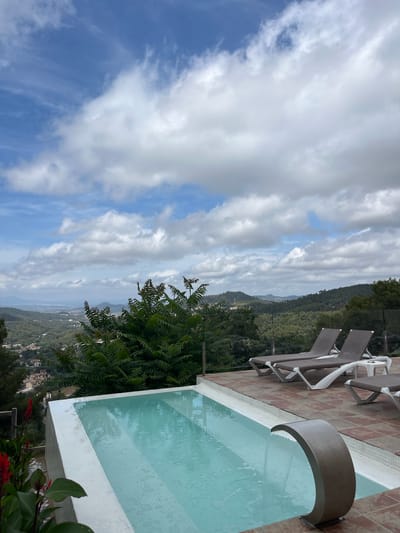 The image depicts an outdoor swimming pool area overlooking a valley with a town and distant mountains. The pool, a rectangular shape with white tiled edges and steps, is filled with clear, pale blue water. A curved, metallic waterfall feature is visible in the foreground of the pool, with water flowing from it. Surrounding the pool is a paved patio area with terracotta-colored tiles. Several lounge chairs, with dark grey cushions and white frames, are arranged on the patio. In the background, a lush green landscape of trees and hills stretches towards a hazy horizon. The sky above is a mix of blue and white clouds. The scene appears to be located in Can Güell, Spain, given the context provided. There are no people visible in the image.