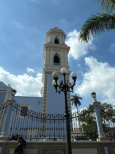 An upward, angled shot captures a tall, blue-and-cream-colored tower against a sky with scattered clouds and a palm frond in the upper left corner. A decorative, wrought-iron fence with pillars runs along the right side, partially obscuring a building behind it. A vintage lamppost with globe lights is centered in the frame. Part of a person is visible in the lower right corner, behind the fence.