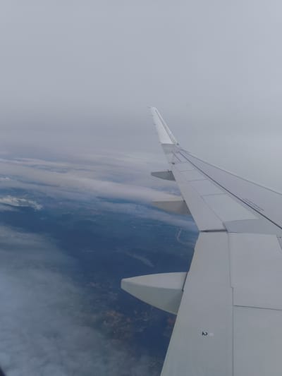 An aerial view captures the wing of an airplane extending into a cloudy sky over Vietnam. The wing, predominantly white, is positioned in the foreground and to the right of the frame. Beneath the clouds, patches of green and brown land are visible, suggesting a rural or semi-rural landscape. A winding river can be seen cutting through the terrain. The sky is overcast with soft, diffused light. The image appears to be taken from a passenger window on a flight.