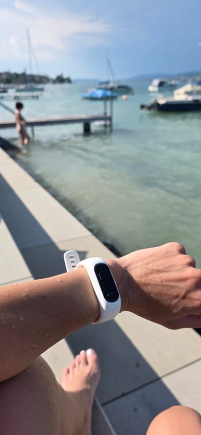 The image is a close-up shot of a person's arm wearing a white fitness tracker, with their feet visible in the foreground. The scene appears to be outdoors, next to a body of water. In the background, a person is standing on a dock, and several boats are visible on the water. The sky is partly cloudy. The water appears to be calm, and there are some shadows on the ground from what looks like a pier or boardwalk. This location is likely the shore of Lake Zurich in Oberrieden, Switzerland, given the presence of boats and the general appearance of the landscape.