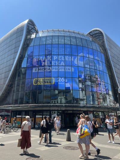 This is a vertical, eye-level, outdoor shot taken on a sunny day, likely in the afternoon, of people walking on a sidewalk in front of a modern, glass-fronted building. The building's facade is dominated by large blue advertisements. One prominent ad features a bottle of "Müllermilch PROTEIN" and text in Polish, including "Z MOCĄ PROTEIN" (With the Power of Protein) and "0% CUKRU" (0% Sugar). Another section of the facade displays text that reads "GALERIA KATOWICKA", indicating the location as a shopping gallery in Katowice, Poland.
Several individuals are visible in the foreground and mid-ground, walking in different directions. A woman in the foreground is carrying a colorful tote bag with a yellow, blue, and red design and is wearing a striped skirt. Another woman in a white top and brown pants is also walking, carrying a brown paper bag. Further back, several other people are walking, some appearing to be tourists or shoppers. The overall mood seems relaxed and casual, typical of a daytime stroll in a city. The reflections on the glass building show the surrounding architecture and the blue sky.