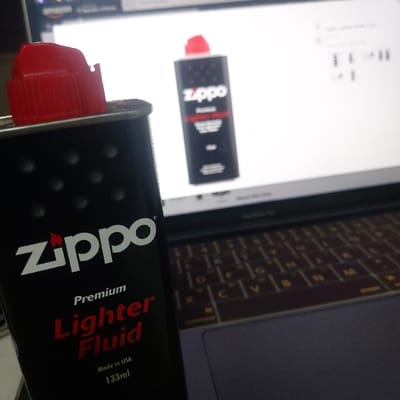 The image displays a close-up, indoor still life scene. In the foreground, a black, rectangular metal can of Zippo Premium Lighter Fluid, 133ml, made in USA, stands upright, prominently featuring its red screw-top cap. The can's label displays the Zippo logo with a flame icon, and "Premium Lighter Fluid" in red lettering. In the background, a laptop with an illuminated screen is visible. The screen displays a blurred image of what appears to be an identical Zippo lighter fluid can on an e-commerce website, with the Amazon logo faintly visible in the upper left corner. The laptop's dark-colored keyboard is partially visible below the screen. No people are present, and no specific actions or interactions are depicted. The scene is illuminated by an indiscernible light source, casting soft reflections. The image contains no discernible visual information to establish a specific location within Gimpo-si, South Korea, or any other geographical area.