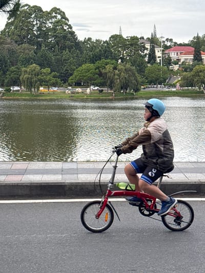In the city of Xuan Huong - Da Lat, Vietnam, a young individual, likely a child, rides a red folding bicycle along a paved road adjacent to Xuan Huong Lake. The cyclist, wearing a blue helmet, glasses, a muted color jacket, and blue cycling shorts with a white logo, is captured in profile, pedaling forward with their right foot pushing down. The bicycle has small wheels and a green water bottle attached to the frame. The background shows the rippling surface of Xuan Huong Lake, a walking path with gray and pink tiles, and a lush green landscape with trees and buildings in the distance. The sky is overcast.