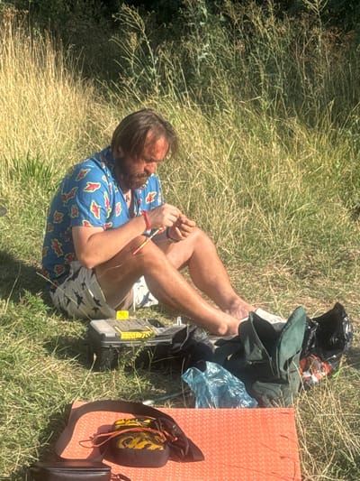 The image depicts a man sitting on the ground in a grassy, outdoor environment, likely in Муромское сельское поселение, Ukraine. He is wearing a blue shirt with a colorful pattern and patterned shorts. The man is focused on an activity involving small objects, possibly crafting or repairing something with his hands, as he holds a string of beads. Around him, there are various items including a fishing tackle box, a dark bag, a green bag, a blue plastic bag, and an orange mat. The background consists of tall dry grass and some foliage, suggesting a rural or park-like setting. The lighting indicates it is daytime, with sunlight casting shadows.