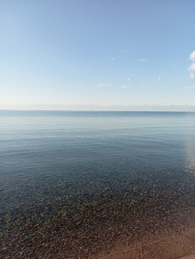 A vertical, eye-level shot captures a tranquil scene at a lakeshore in Bosteri, Kyrgyzstan. The image is divided into three horizontal segments: a vast expanse of clear blue sky with wispy white clouds occupying the top third, a calm, rippling body of deep blue water filling the middle section, and the bottom third showing a clear view of a pebble-strewn lakebed under shallow, crystal-clear water, with a sandy shoreline visible at the very bottom right edge. The distant horizon is marked by a thin, flat line of snow-capped mountains. No people or man-made objects are present. The lighting suggests a clear day, possibly late morning or early afternoon.