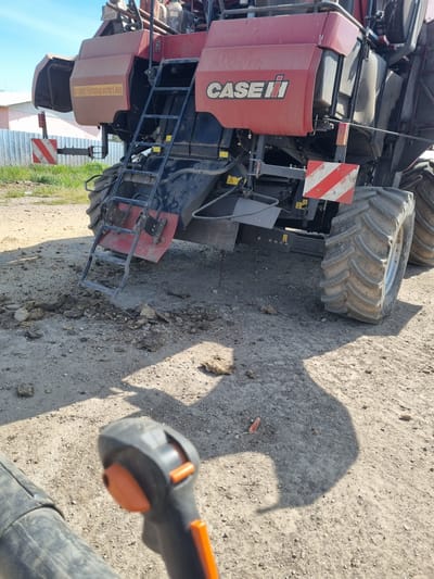 A large red and black combine harvester, branded "CASE IH," is parked on an unpaved, dusty, and rocky surface, likely a farmyard or rural road in Ovcharovo, Bulgaria. The machine is equipped with massive, heavily treaded tires and a black metal ladder for access. A red and white striped warning sign, typical for oversized agricultural vehicles, is affixed to its side.
In the lower right foreground, the orange and black handle of a leaf blower or similar cleaning device is partially visible, indicating a potential cleaning or maintenance activity. A distinct shadow, likely cast by a person or object, stretches across the dusty ground, suggesting bright sunlight. The ground is littered with small rocks and dried earth debris.
The scene is set outdoors under clear, sunny skies, with strong shadows indicating either morning or late afternoon. In the background, a light-colored fence or building structure can be partially seen.
Visible text includes the "CASE IH" logo on the side of the harvester. A yellow warning sticker on the machine's side reads "ACHTUNG! FAHRZEUG SCHERT AUS," which is German for "CAUTION! VEHICLE SWINGS OUT."