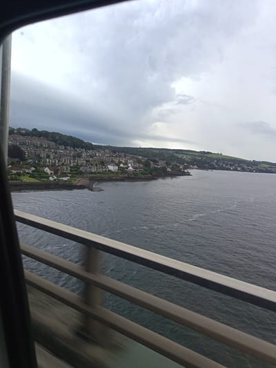 The image captures a panoramic view of a coastal town from a slightly elevated perspective, likely from a bridge or a high vantage point. In the foreground, a weathered metal railing with horizontal bars stretches across the frame, indicating the viewpoint is from a structure overlooking the scene. The railing is a prominent object in the image.
The main subject is the town and the body of water it borders. The town is nestled along a hilly coastline, characterized by a dense arrangement of houses with pitched roofs, mostly in muted tones of grey, white, and brown. The architecture suggests a residential area, possibly a suburb or a part of a larger city. Green trees and foliage cover the hills behind the houses, creating a contrast with the urban sprawl.
The body of water is a large expanse of dark, choppy sea, with ripples and white streaks suggesting moderate movement. The water extends to the horizon, where it meets a dramatic sky filled with heavy, grey clouds. The overcast conditions suggest it is likely a cloudy or potentially rainy day, possibly late afternoon or early morning, given the diffused light.
The setting is clearly coastal, with the town built close to the water's edge. The rocky shoreline is visible in places where the town meets the sea. The location context provided is Dundee, United Kingdom, which is consistent with the appearance of a Scottish coastal town. There is no discernible activity or event taking place, the scene appears to be a static view of the town and its surroundings. No people are visible, and no text is present in the image. The overall mood evoked is one of a slightly somber, perhaps tranquil, but uninspiring day in a picturesque coastal setting.
