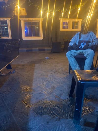 The scene depicts an outdoor seating area at night in Igbessa, Nigeria. A dark-colored building wall serves as the background, featuring two distinct windows with prominent white decorative frames. Above the left window, a "Gree" brand air conditioning unit is mounted. String lights with warm yellow bulbs are strung across the upper portion of the frame, accompanied by artificial green foliage. The lower section of the wall exhibits a blue and white striped pattern near the base.
The ground consists of dark-colored, textured paving, resembling stamped concrete with wood grain and various abstract patterns. Sections of the ground are visibly wet, reflecting the overhead lights. Discarded clear plastic bottles are present on the ground: one centrally located and another near the base of a dark table on the left.
In the mid-ground, a lone individual is seated in a dark, possibly wicker, chair. The person wears a light-colored long-sleeved sweatshirt and matching light-colored sweatpants featuring padded or textured knee areas. Their face is largely obscured by shadow, but dark hair or dreadlocks are visible cascading over their right shoulder. The individual holds a dark bag with a prominent white "NY" logo on their lap, with hands clasped over it. To the right of the seated individual, a black plastic stool is partially visible in the foreground. In the background, a small dark table with chairs is positioned. A blue light emanates from within the left window.