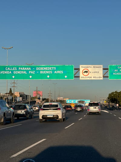 The image shows a multi-lane highway in Boulogne Sur Mer, Argentina. Multiple cars are visible driving on the highway. Overhead signs indicate directions to Calles Parana, Avenida General Paz, Debenedetti, and Buenos Aires. A separate sign indicates "Transito Pesado" (Heavy Traffic) and "V. Lopez" (Vicente Lopez) and "Solo por Pelliza" (Only by Pelliza). The sky is clear.