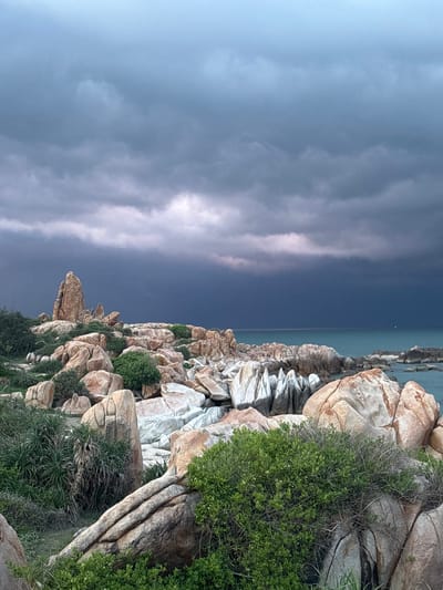 The image depicts a rocky coastline under a dramatic, overcast sky. The foreground is filled with large, weathered boulders of various shapes and sizes, interspersed with hardy green vegetation. The rocks exhibit a mix of textures and colors, ranging from light tan and white to darker earthy tones, some showing signs of erosion and deep fissures. In the mid-ground, the rocky terrain continues towards the sea, with a prominent tall, pointed rock formation standing out against the horizon. The sea is visible in the background, a deep teal color under the heavy clouds. The sky is filled with dark, turbulent storm clouds, with a lighter, diffused light breaking through on the left side, suggesting either the early stages of a storm or its aftermath. The scene appears to be located near a coastal area, possibly within or near the city of Thuận Thành, Vietnam, known for its natural coastal landscapes. There are no people or animals visible in the image.