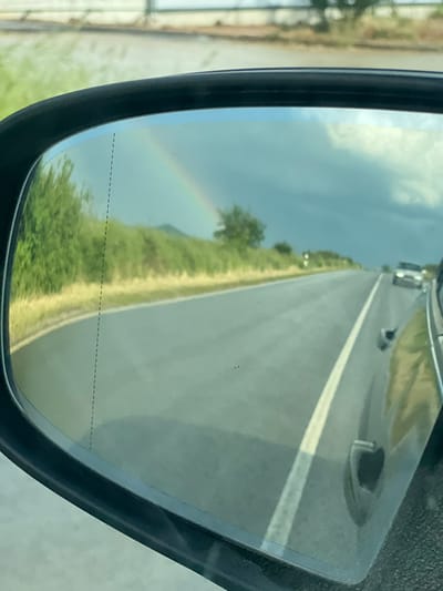 The image shows the reflection in a car's side mirror. The reflection depicts a road, grass, and a rainbow in the sky. Several vehicles are seen on the road. The dashed line on the side mirror indicates that the mirror has blind spot monitoring. It is not possible to identify a specific location in the city of Brezovo, Bulgaria from the details available in the image.