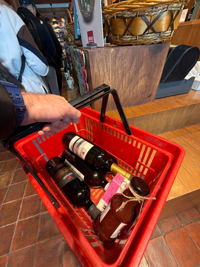A high-angle view shows a person holding a red plastic shopping basket filled with three bottles of wine and a bottle of sauce, positioned on a tiled floor. The person's hand and a portion of their arm wearing a blue watch are visible holding the black handle of the basket. The basket contains three wine bottles and a sauce bottle. The wine labels are visible, with the text “Winery Khareba” and “Wine of Georgia” appearing on them. The basket is red. The tiled floor consists of reddish-brown tiles. In the background, there are items on shelves, including wooden baskets, boxes, and other products typically found in a store. The store contains items such as souvenirs, alcohol, and other goods. The scene suggests a retail environment, possibly a wine shop in Gamarjveba, Georgia.