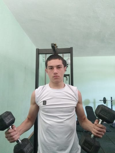 A young man stands in a gym in Gradets, Bulgaria, holding two 10 kg dumbbells. He is wearing a white sleeveless t-shirt. Behind him is a weight machine and in the background, a weight bench and barbell are visible. The walls are pale green.