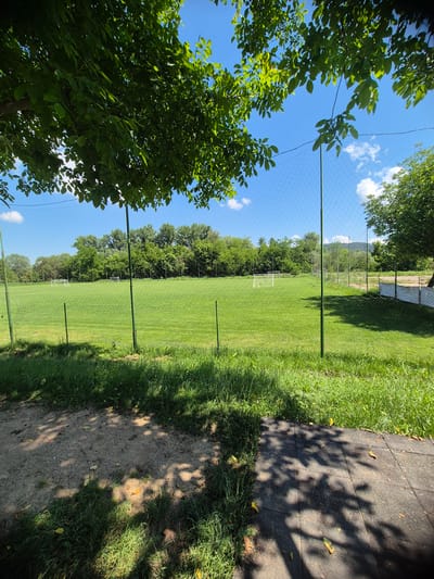 This is a medium-shot, eye-level, outdoor photograph taken on a sunny day in the town of Debelets, Bulgaria. The foreground shows a patch of sandy ground with green grass and dappled sunlight from overhanging tree leaves. A dark gray paved walkway runs along the bottom right edge of the frame. A green net fence, supported by thin green poles, separates the foreground from a large, vibrant green soccer field. In the distance, on the soccer field, are two white soccer goals. The field is surrounded by a line of lush green trees. The sky is a clear, bright blue with a few scattered white clouds. The overall impression is of a sports field on a clear, pleasant day.