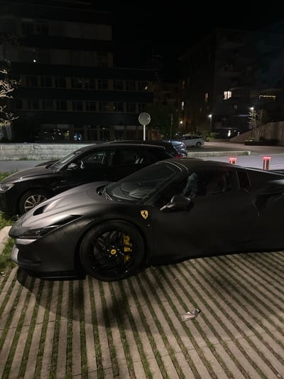 The image displays a dark-colored Ferrari sports car, likely matte black or dark grey, parked at night in an urban or suburban setting in Dübendorf, Switzerland. The car is positioned on a paved surface composed of striped concrete pavers with grass growing in the gaps. Its black multi-spoke wheels showcase bright yellow brake calipers and a yellow Ferrari emblem on the wheel hub, matching a larger emblem on the front fender.
In the immediate vicinity of the Ferrari, a clear plastic bottle with unreadable white and red text lies on the ground. Another lighter-colored car is partially visible behind the Ferrari. The background is dominated by several dark, multi-story buildings, some with illuminated windows, suggesting residential or office blocks. Streetlights and red-glowing bollards provide additional artificial illumination. A small, round mirror on a pole is visible on the far left. The sky is dark, indicating nighttime, and no people or specific activities are taking place; the cars are stationary. The scene appears dry and still.