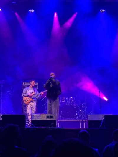The image depicts a live musical performance, likely a concert, taking place in an indoor or nighttime outdoor venue characterized by heavy artificial lighting and atmospheric haze. Two male musicians are prominent. One man stands wearing sunglasses and a dark jacket, possibly a singer or performer. The other man is holding a bass guitar, wearing a light-colored shirt with "GR" printed on it, presumably playing it. They are standing on a stage with amplifiers. There are drums and other musical equipment visible behind them. Lighting consists of blue and pink spotlights and a multi-bulb fixture. Silhouettes of audience members are seen in the lower right corner. Although the image does not provide information that will definitively identify Vilanova i la Geltrú, Spain, there is no information in the image that would exclude it from being in a city in Spain.