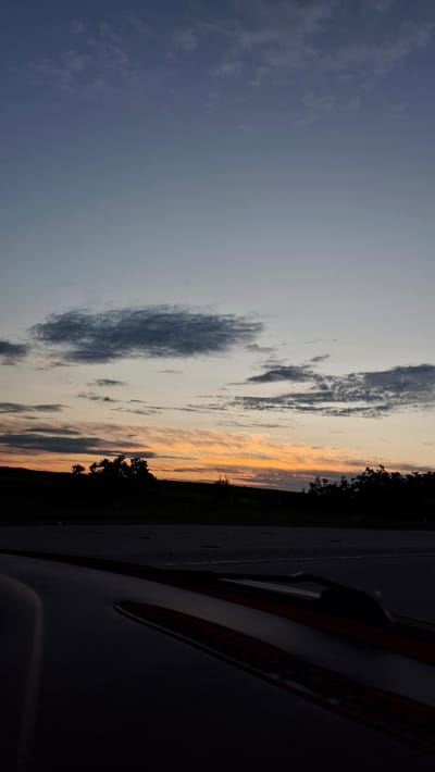 The image depicts a dusky sky over a landscape of rolling hills and silhouetted trees. The sky transitions from a pale blue at the top to shades of orange and yellow near the horizon, indicating either sunrise or sunset. Dark, wispy clouds are scattered across the sky. In the foreground, the dark, reflective surface of a vehicle is visible, with some orange accents. The scene appears to be a roadside view. The location is identified as Razeni, Moldova.