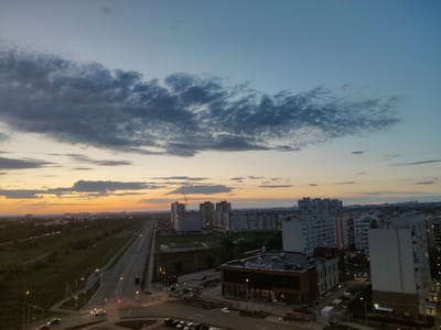 This aerial shot captures a dusky twilight scene over a suburban or semi-urban landscape in Придорожный, Russia. The sky is a gradient of soft blues and oranges, with scattered, dark, and wispy clouds near the horizon, indicating the end of daylight. The setting appears to be a developing residential area, with numerous apartment buildings of varying heights and architectural styles dominating the mid-ground and background. Many of these buildings are illuminated, suggesting that it's evening.
In the foreground, a multi-lane road stretches into the distance, with a few cars and trucks visible, their headlights and taillights faintly glowing. Streetlights line the sides of the road and some intersections. Adjacent to the road, there are patches of green, likely undeveloped land or parks, and some scattered smaller structures. One notable building in the mid-ground has prominent signage with Russian text, "СОЛНЕЧНЫЙ" (Solnechnyy), meaning "Sunny," which contrasts with the darkening sky. A construction crane is also visible in the distance, hinting at ongoing development. The overall mood is calm and transitional, transitioning from day to night. There are no people clearly visible in the image.