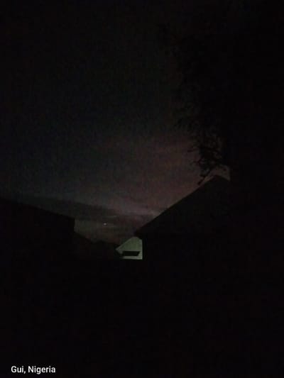 The image is a dark, nighttime photograph taken in Kuje, Nigeria. Silhouetted against a dimly lit sky, the outlines of several buildings are visible. One building in the foreground, on the right, has a sharply angled roof. In the distance, a faint light is visible, possibly from a distant town or structure. The sky itself is cloudy and appears to have a reddish hue in some areas, suggesting a twilight or possibly atmospheric phenomenon. The lower left corner of the image contains text that reads "Gui, Nigeria". The overall impression is of a quiet, rural or suburban night scene.