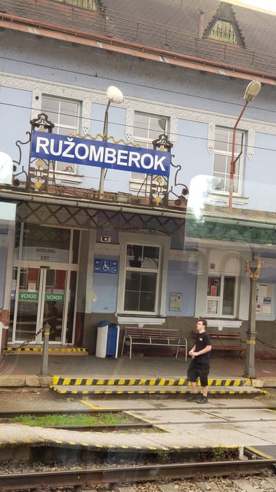 The image shows the exterior of the Ružomberok train station in Slovakia. A blue sign above the entrance reads "RUŽOMBEROK". The building is light blue with decorative white trim. There are several windows and doors. To the left of the entrance, there are signs indicating "VCHOD", "AUSGANG", and "EXIT". To the right of the entrance, there are signs for "WC" and a wheelchair accessibility symbol. A young man in a black t-shirt and shorts stands on the platform, holding a cup. Benches are visible on the platform. The platform has yellow and black striped safety markings. Train tracks are in the foreground.