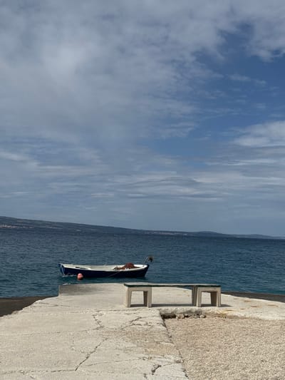 The image shows a concrete pier in Dugi Rat, Croatia (43.446, 16.634). A small blue and white boat is docked in the water next to the pier, and a bench sits atop the pier. There is a faint line of land in the distance. The sky is a clear blue with wispy white clouds. The water appears calm and peaceful, with a hint of a wave in the foreground. The scene is deserted, with no people present.