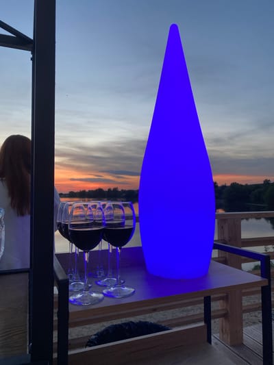 Here is a forensic description of the image:
**Scene:** Outdoor setting, likely a terrace or balcony. Background shows a sunset-lit sky over a body of water bordered by trees.
**Objects:** A luminous, blue, elongated object on a wooden table, wine glasses, a dark metal frame, wooden structures, a person with reddish-brown hair, and crumpled foil.
**Details:** The elongated object appears to be a light fixture, providing illumination on the table. There are four wine glasses, suggesting an intention for use. The metal frame and wooden structures indicate the presence of architectural elements. The reddish-brown hair suggests a person seated or present nearby. The crumpled foil might indicate food or remnants.
**Environment:** The sky showcases colors associated with sunset. Vegetation is visible across the body of water.