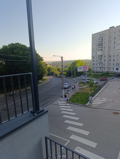 This image captures an outdoor scene in Fastiv, Ukraine, likely during the late afternoon or early evening, given the warm, soft light and long shadows. The setting appears to be a residential area on the edge of a town or city.
In the foreground, a dark metal railing and a textured grey wall suggest the viewpoint is from a balcony or elevated structure. Beyond this, a paved area with white crosswalk markings is visible, indicating a pedestrian crossing. A barrier arm, common in parking areas or restricted zones, is present at the edge of the pavement.
Further into the scene, a road curves through a somewhat hilly landscape dotted with trees. On the left side of the road, a dark-colored motorcycle is parked near a metal fence. Several people are walking on the sidewalk or crossing the street. Among them are two women walking together, and another woman walking away from the camera across the crosswalk. A few cars are parked on the side of the road and in a designated parking lot on the right.
A large, multi-story apartment building, characteristic of Eastern European architecture, dominates the right side of the frame. Its facade is light-colored brick with numerous windows and balconies. In the background, a bridge spans a distant valley, and beyond that, more trees and a few scattered buildings can be seen, suggesting a larger urban area or the outskirts of the town.
A prominent billboard stands near the road, featuring pink and white branding and text that appears to be in Cyrillic. The sky is a clear, pale blue, with the sun setting behind the distant horizon, casting a warm glow. The overall atmosphere is calm and ordinary, depicting daily life in a Ukrainian town.