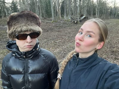 The image depicts two individuals, a male and a female, in an outdoor setting in Djurhamn, Sweden, likely captured as a selfie. The male, positioned on the left, is wearing a brown fur-like aviator hat and rectangular, amber-tinted sunglasses. He has a serious, unsmiling expression and is dressed in a shiny black hooded puffer jacket with silver-colored hardware. The female, positioned on the right and appearing to hold the camera, has long blonde hair styled in a braid over her left shoulder, secured with a decorative hair tie. She wears small gold hoop earrings and a dark-colored zippered jacket. Her facial expression is a pouting "kissing" pose. The background features a sparse deciduous forest with bare trees and fallen leaves covering the ground, indicating a late autumn or winter season. A pile of cut logs or branches is visible in the middle ground, suggesting recent clearing activity.