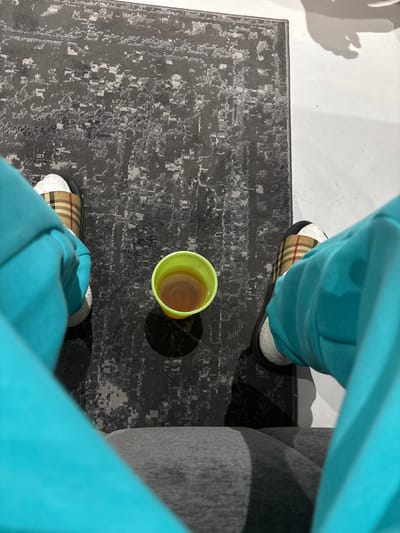 An overhead, first-person perspective image depicts an indoor scene in Hartebeespoort, South Africa. The lower half of a person's body is visible, with the lap covered by grey fabric and the upper legs clad in bright turquoise/teal track pants. Both feet, positioned on a dark grey area rug with a distressed light grey pattern, are wearing slider-style slippers. Each slipper features a white terry cloth lining, a dark sole, and a strap patterned with a tan, red, and black plaid design. Centered between the feet on the rug is a lime green plastic cup containing a translucent amber-brown liquid. A distinct circular shadow is cast directly beneath the cup on the rug. To the right, a section of a white hard floor is visible adjacent to the rug, exhibiting a faint, dark, irregularly shaped shadow.