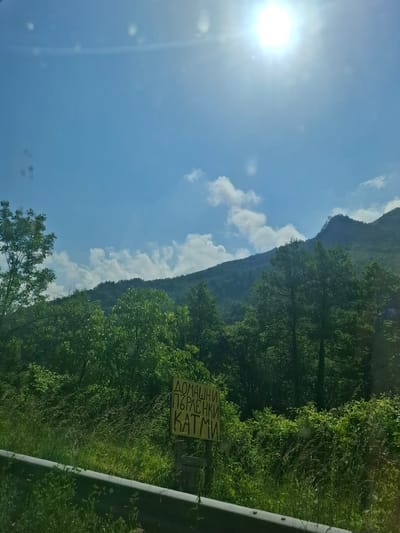 This image captures a bright, sunny daytime landscape, likely taken from a moving vehicle near Нареченски бани, Bulgaria. The sun, prominent with strong lens flare, illuminates a clear blue sky dotted with scattered white clouds.
The scene is dominated by a majestic, tree-covered mountain range in the background, showcasing dense green foliage. In the midground and foreground, lush green trees and undergrowth line what appears to be a roadside. A grey metallic guardrail or barrier runs vertically along the right side of the frame, confirming the presence of a road.
A notable object is a yellow rectangular sign situated amidst the greenery on the right. The sign bears Cyrillic text, which reads:
ДОМАШНИ
ПЪРЛЕНКИ
КАТМИ
This translates to "Homemade Părlenki Katmi," indicating that traditional Bulgarian flatbreads (părlenki) and crepes/pancakes (katmi) are being offered for sale, likely by a local vendor along the road. The overall impression is one of a scenic natural environment on a pleasant day, with a local culinary touch.