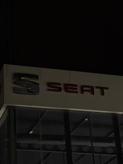 The image displays the upper section of a commercial building's facade at night, located in Polígon Industrial De Son Castello, Spain. A prominent "SEAT" corporate logo is mounted centrally on a light-colored, possibly marble-patterned, horizontal panel. The stylized 'S' is a metallic, chrome-finished emblem, while the letters "SEAT" are red and appear to be illuminated. Below the logo, a large dark glass window reflects a dimly lit interior space. Visible within this reflection are dark horizontal slat-like structures, likely part of a ceiling or internal blinds, along with faint interior lighting. A small, circular, light-colored object, consistent with a smoke detector or recessed light, is discernible on the reflected ceiling towards the right. The sky above the building is uniformly dark. No individuals or specific actions are visible.