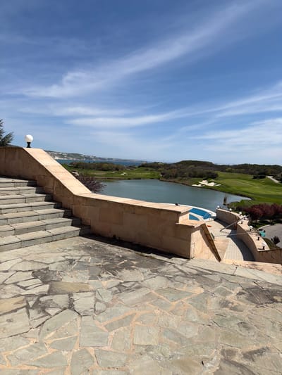 The image, rotated to its correct horizontal orientation, presents an elevated panoramic view of a scenic coastal landscape in Bozhurets, Bulgaria. In the foreground, on the right, a textured stone pavement with irregular, flat gray and brown flagstones leads down a set of light-colored stone steps. A matching stone wall or balustrade runs alongside these steps, featuring a spherical white lamp post at its base.

Further into the midground, integrated into the stone architecture, is a small, inviting blue swimming pool with steps. Beyond this structure lies a serene body of water, likely an inlet or bay of the Black Sea, stretching horizontally across the scene. The opposite bank features a vibrant green golf course with manicured fairways, scattered trees, and prominent white sand bunkers. Distant land formations or cliffs are visible along the horizon on the far side of the water.

The background is a vast, clear blue sky adorned with elegant, wispy white clouds, indicative of a bright, sunny day. The strong sunlight casts distinct shadows, suggesting the time of day is late morning or early afternoon. The overall setting is tranquil and picturesque, highlighting a resort-like environment. No people or visible text are present in the image.