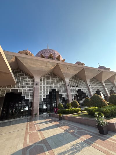 This is a low-angle, wide shot of the Putra Mosque in Putrajaya, Malaysia. The mosque, built on the edge of Putrajaya Lake, features a pink dome with intricate white patterns and is surrounded by manicured bushes and trees. The foreground consists of a wide paved walkway with a geometric pattern, and the building's exterior is characterized by its large, arched windows with decorative latticework and its prominent triangular architectural elements. The sky is clear and blue, indicating a sunny day. Two individuals in red traditional clothing can be seen near the entrance of the mosque.
