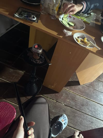 The image depicts an outdoor, low-light setting with a wooden floor and a wooden table. On the table, there are several plates with food, including sliced cucumbers and what appears to be potatoes. Utensils and napkins are also present. A person's hand is seen reaching for a cucumber slice. To the left of the table, a hookah with glowing coals is visible. In the foreground, a person's legs and feet are in view, with one foot wearing a grey Croc shoe adorned with colorful charms. Another person's arm wearing a jacket with red and blue stripes is also visible. The environment suggests a gathering or meal taking place outdoors, possibly in Zhebrei, Russia, given the context.